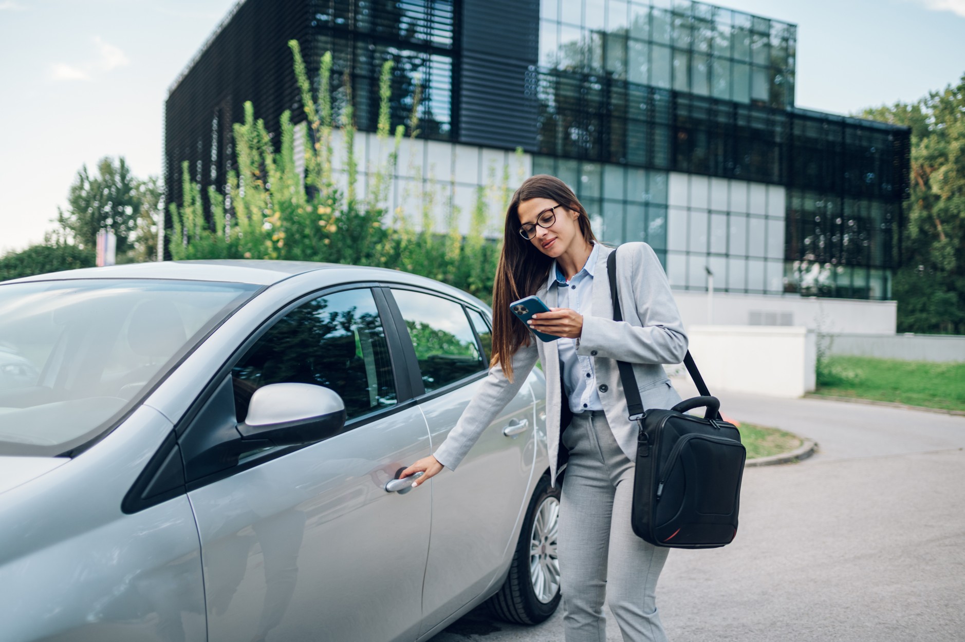Businesswoman in a suit unlocking a silver company car outside an office, illustrating business car insurance in South Africa.